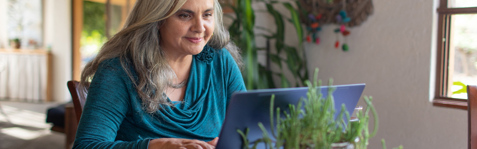 Older woman sat at her table with plants around her on her laptop, working on her online community engagement volunteer role.